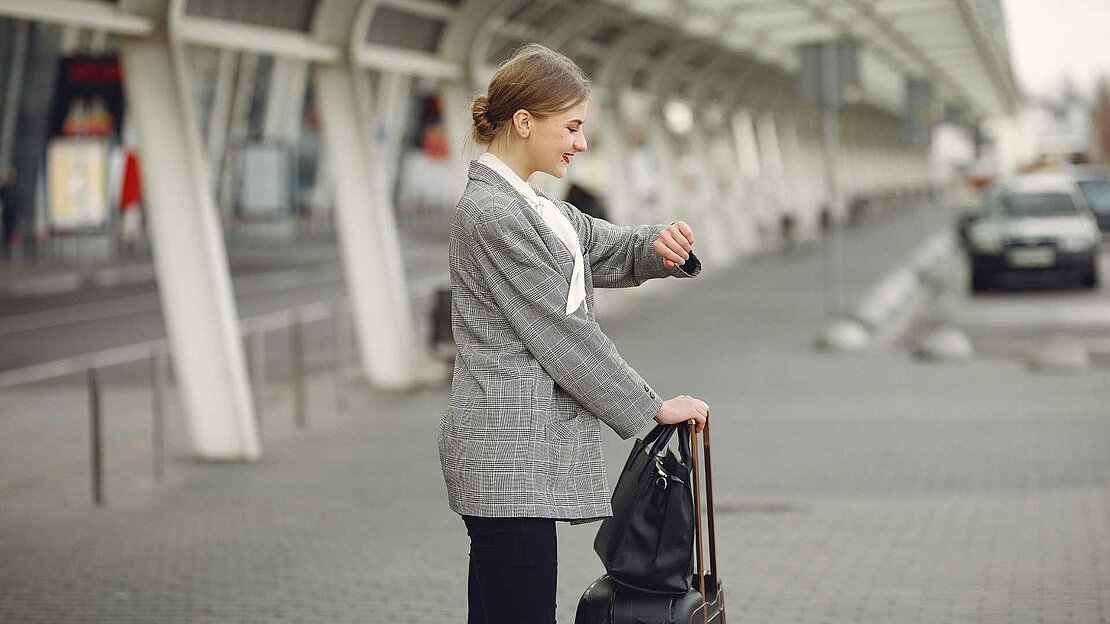 Foto: Frau steht mit Koffer am Flughafen und schaut auf ihre Armbanduhr